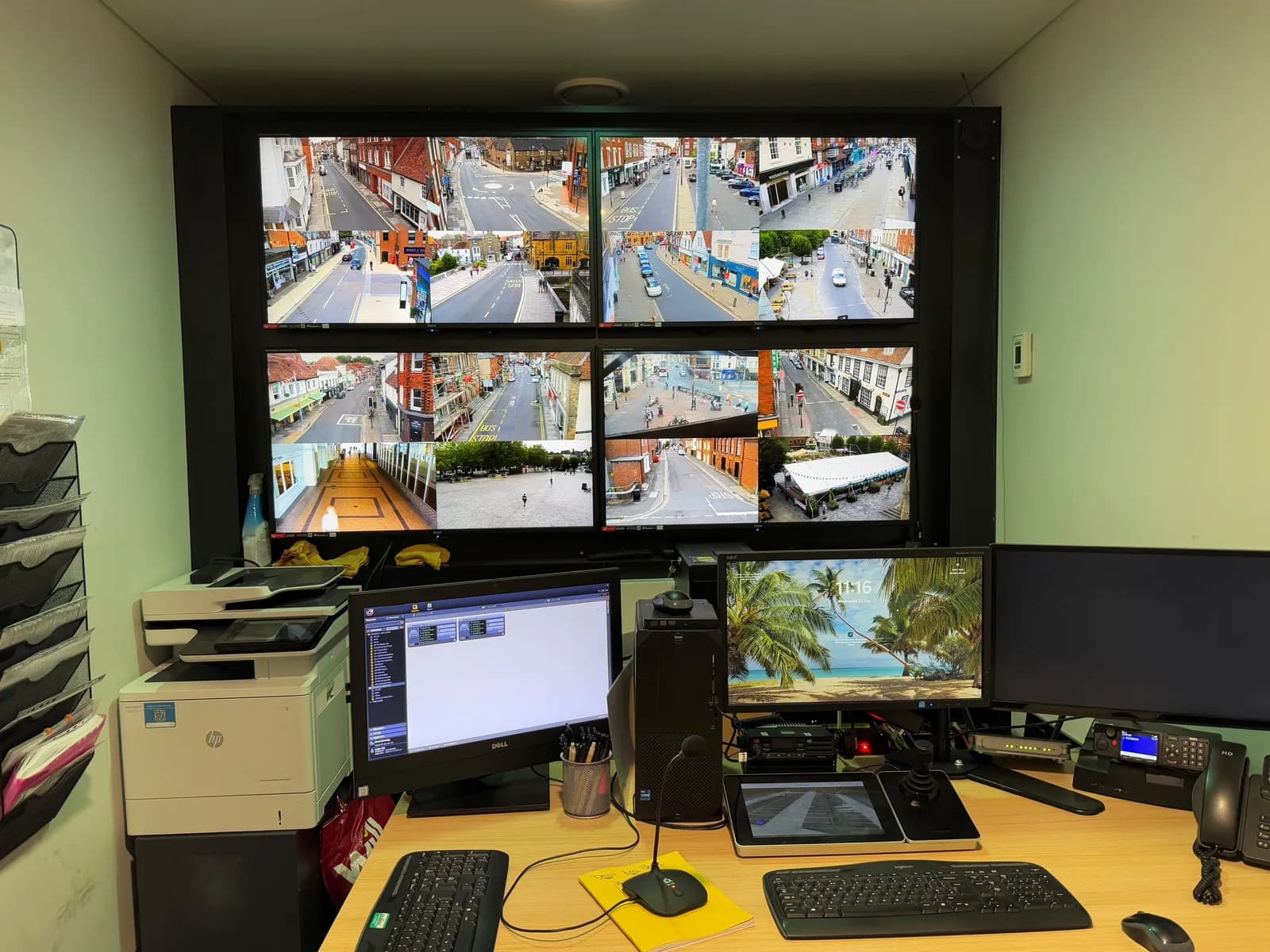 A control room desk with multiple computer monitors, keyboards, a printer, and a large display wall showing several live camera feeds of city streets and public spaces.